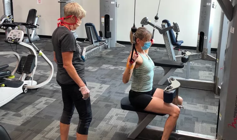 a personal trainer in a mask and gloves watching participant with mask on use a pull-down machine while socially distanced