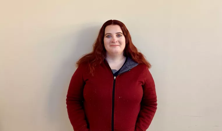 Beth, a white woman with red hair wearing a red zip up sweater, stands in front of a white wall and smirks at the camera