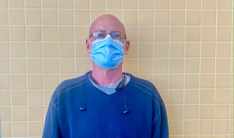 A man in a blue sweater and blue disposable face covering stands in front of a tan-tiled wall while looking at the camera