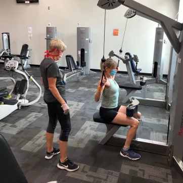 a personal trainer in a mask and gloves watching participant with mask on use a pull-down machine while socially distanced
